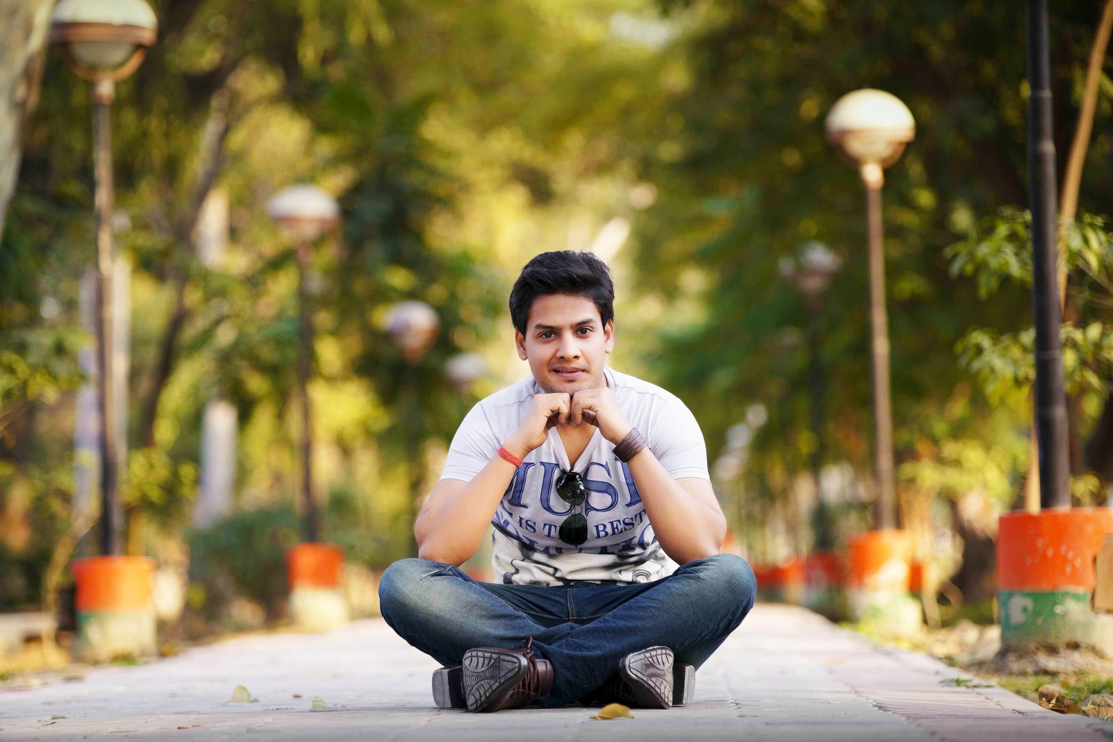 Stylish young man sitting cross-legged on a park path with lamps lining the way.